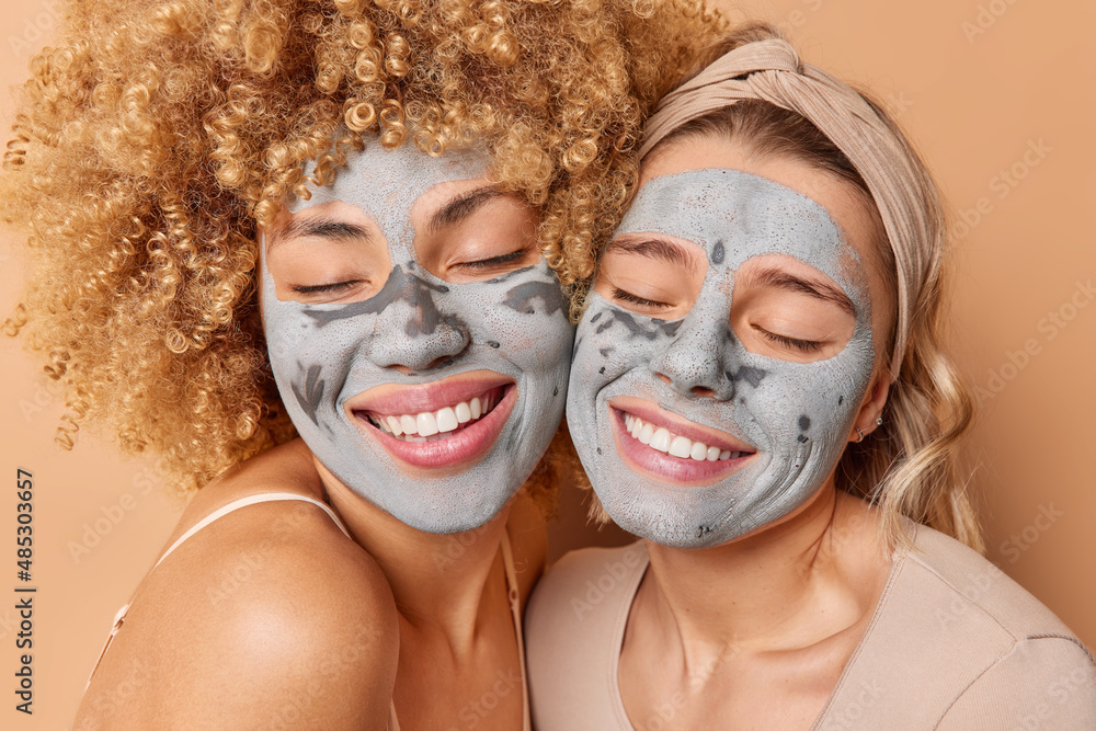 Happy women with clay masks on faces smile happily keep eyes closed from satisfaction take care of skin isolated over brown background express positive emotions. Wellness and beauty concept.