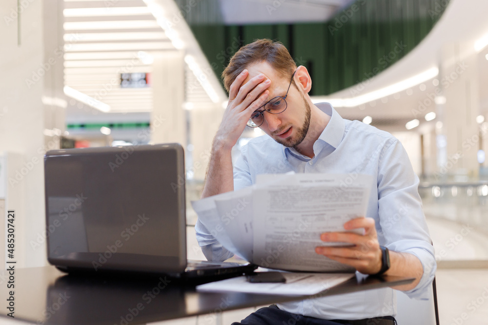 Young puzzled businessman working in empty food court in mall with documents and laptop Young puzzled businessman working in empty food court in mall with documents and laptop