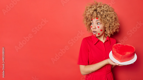 Shocked woman with curly bushy hair holds delicious cake prepared for Valentines Day focused away wears dress poses against red background blank space for your advertising content watches her love