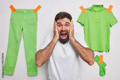 Bearded emotive Caucasian man grabs face keeps mouth widely oened reacts on something amazing poses against white background green trousers t shirt and sock plastered to wall. Human reactions photo