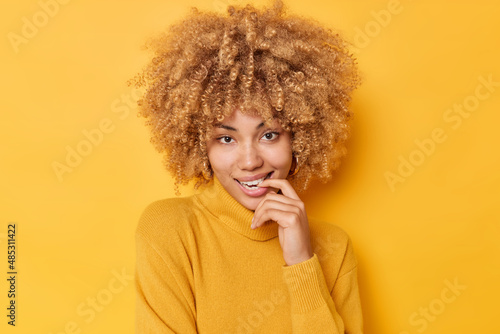 Flirty charming lovely woman bites finger looks directly at camera ha fair curly bushy hair wears casual jumper isolated over vivid yellow background. People facial expressions and dreams concept