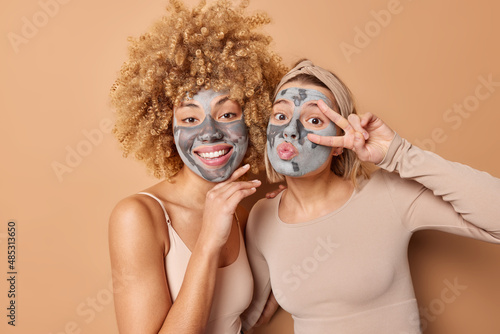 Happy friendly two women pose indoor with facial clay masks smile and make peace gesture over eye take care of skin and complexion dressed casually isolated over brown background. Beauty time © wayhome.studio 