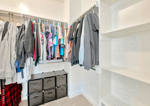 Small walk-in closet with hanging sweaters, shirts and blouse on the metal rods © Jason