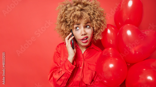 Unhappy crying woman answers phone call upset nobody congratulated her with Valentines Day wears stylish shirt holds heart shaped helium balloons isolated over red background with copy space