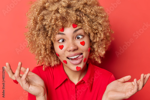 Funny curly haired blonde young woman with sticky hearts on face prepares for St Valentiines Day makes grimace crosses eyes sticks out tongue spreads palms isolated over vivid red background photo