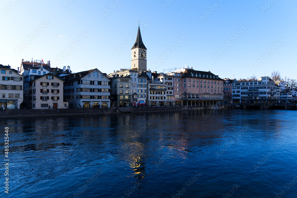 Church tower of medieval protestant church St. Peter at the old town of Zürich with river Limmat in the foreground on a sunny winter day. Photo taken February 5th, 2022, Zurich, Switzerland.