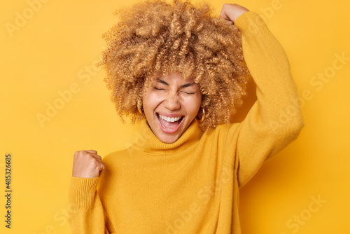 Valokuva Overjoyed curly haired woman makes yes gesture feels excited and triumphant exclaims from joy wears casual sweater poses over yellow studio background celebrates success