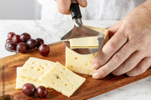 Male hands cutting a piece of cheese with special slicer on wooden board on marble table in the kitchen. photo