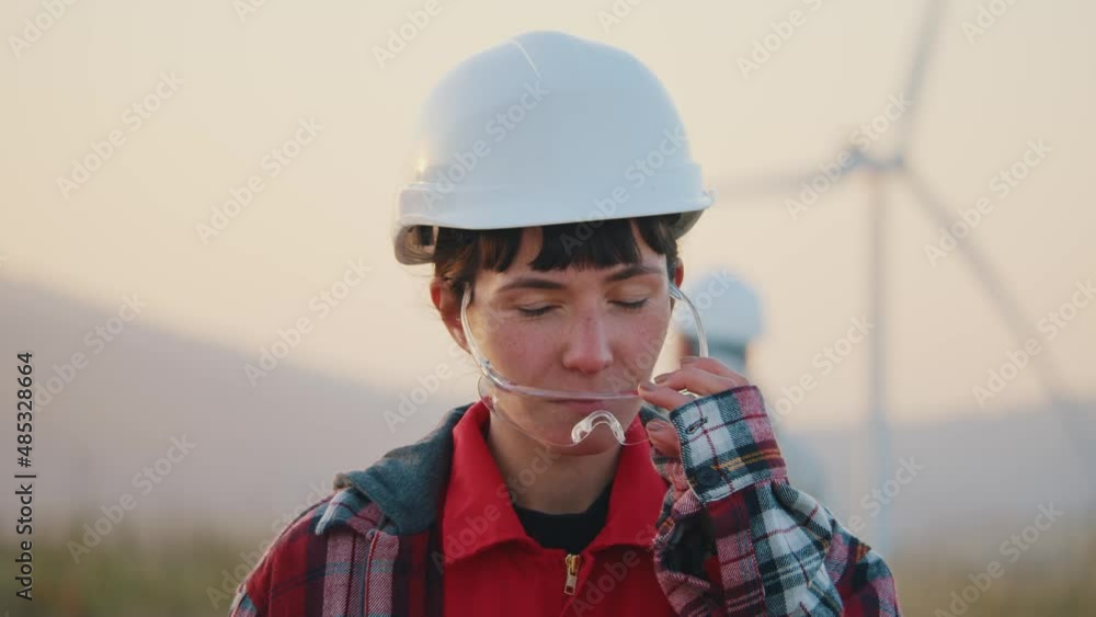Attractive portrait of young woman engineer in a white helmet look at camera removes goggles stand on ecological station. Wind turbines rotating at sunset. Slow motion Attractive portrait of young woman engineer in a white helmet look at camera removes goggles stand on ecological station. Wind turbines rotating at sunset. Slow motion