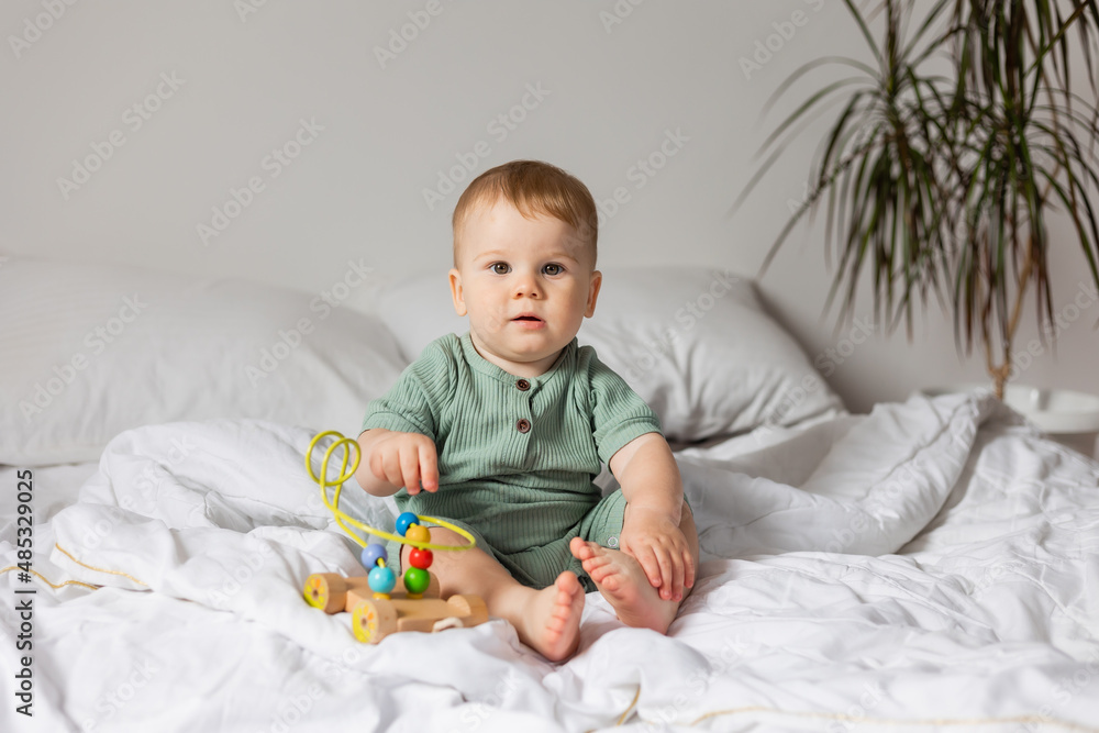 cute baby in a green cotton bodysuit plays with wooden educational toys while sitting in bed. products for children, early development, nursery kindergarten. space for text. High quality photo cute baby in a green cotton bodysuit plays with wooden educational toys while sitting in bed. products for children, early development, nursery kindergarten. space for text. High quality photo