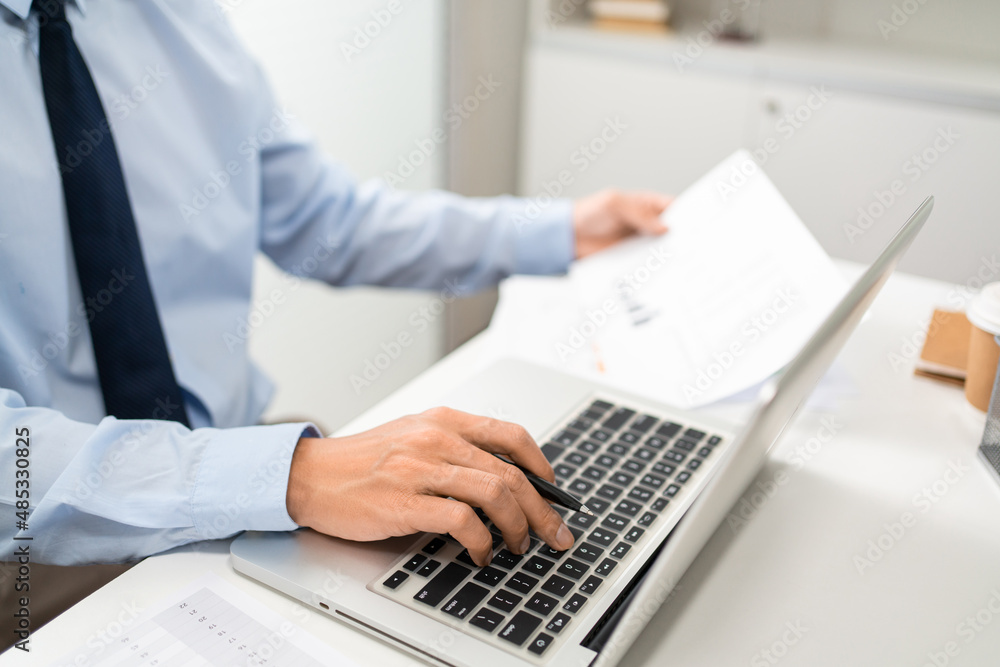 Working Man Conept The man sitting in front of the computer laptop at his desk, holding the document with his left hand and using his right hand holding a pen Working Man Conept The man sitting in front of the computer laptop at his desk, holding the document with his left hand and using his right hand holding a pen
