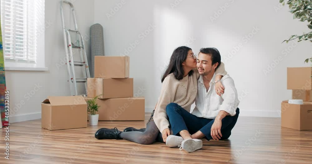 Happy contented young couple moves into their first apartment together, couple sits on a wooden floor close together, an empty room, boxes of unpacked stuff strewn everywhere, freshly renovated Happy contented young couple moves into their first apartment together, couple sits on a wooden floor close together, an empty room, boxes of unpacked stuff strewn everywhere, freshly renovated
