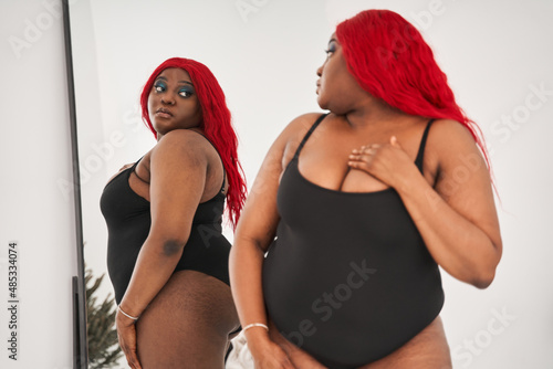 Red haired overweight young woman in black underwear standing in front of the mirror photo