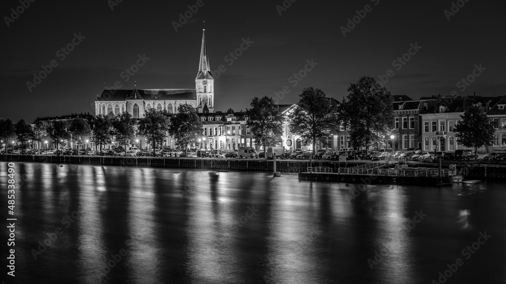 An illuminated church towers from the skyline along a river in the city of Kampen in the Netherlands
