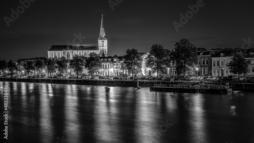 An illuminated church towers from the skyline along a river in the city of Kampen in the Netherlands