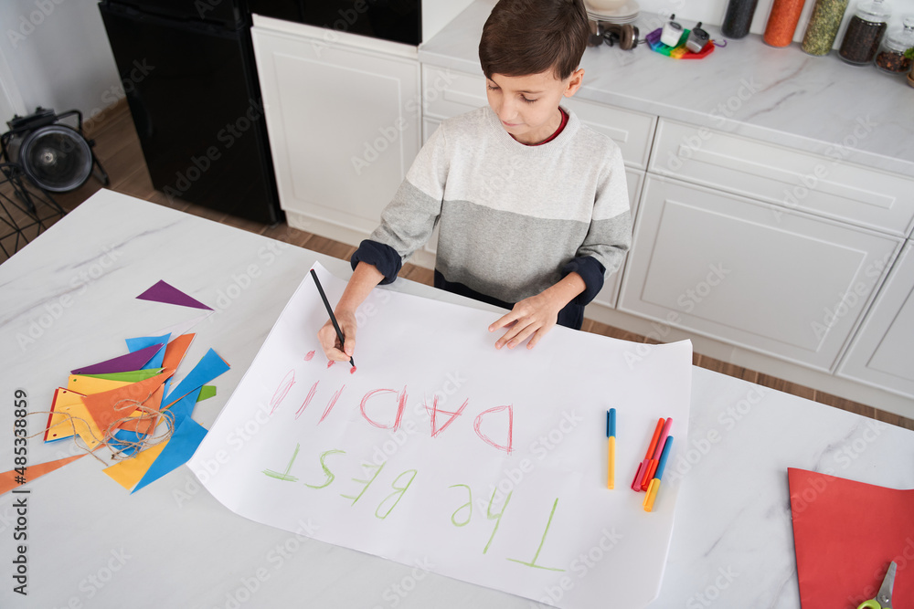 Caucasian boy sitting at the table and drawing placard for his father at home Caucasian boy sitting at the table and drawing placard for his father at home