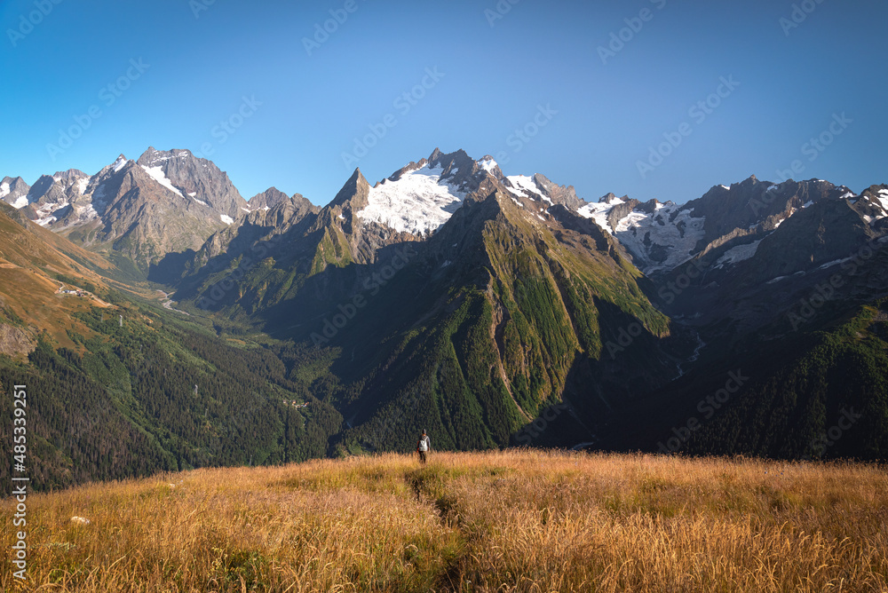 Mountaintops of Dombai in summer with a meadow and a man in the distance