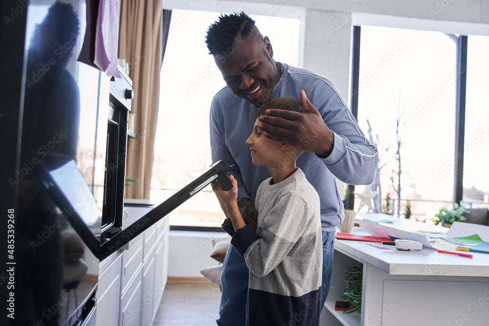 Fototapeta premium Happy multiracial man bonding to his curious caucasian son while he looking into the oven