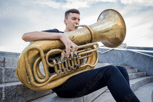 Young street musician playing tuba sitting on granite steps