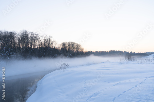 Fog under river and frozen trees on the snowy bank.