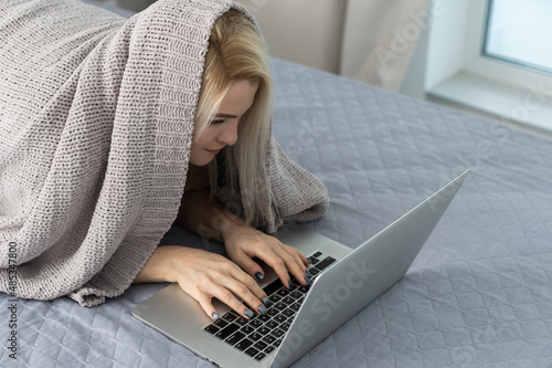 Woman looks with interest at a laptop, hiding under a blanket, in bed, photo