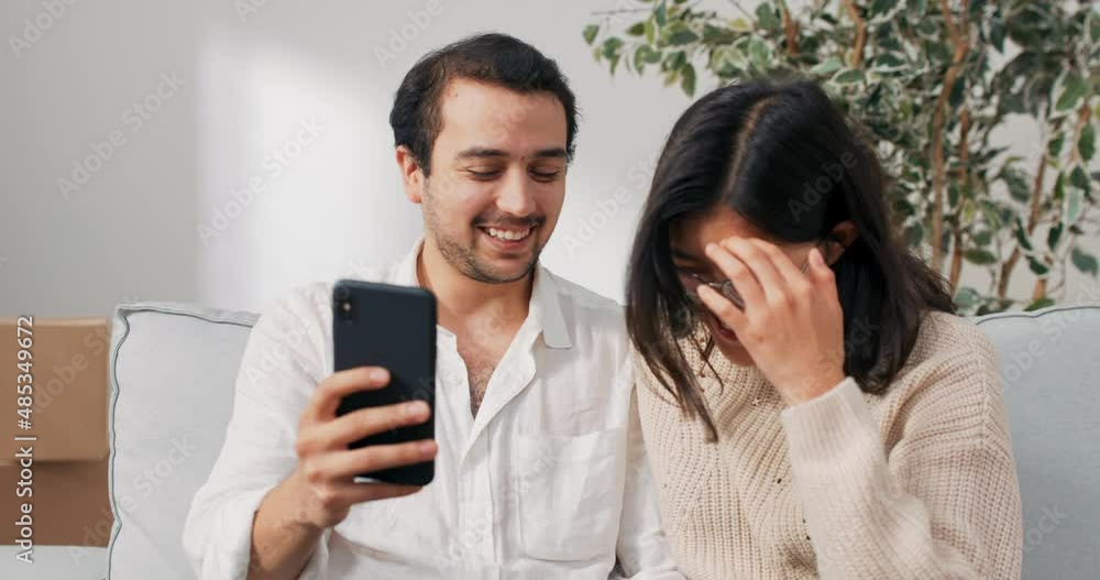 Young couple in love spending time in their new apartment are sitting on the couch looking at the phone, looking for furniture on the Internet, decorations, thinking about the choice of inspiration