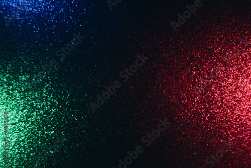 Color glow background. Blur glitter texture. Party illumination. Defocused neon green red blue shimmering sparkles pattern on dark black abstract overlay.