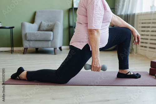 Lower part of active senior woman holding dumbbell during physical exercise on yoga mat on the floor of living-room at leisure
