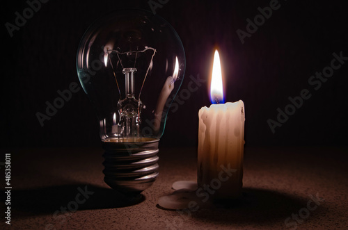 Switched off light or not glowing light bulb near a burning candle in complete darkness. Blackout city, electricity off, energy crisis or power outage, concept image.
