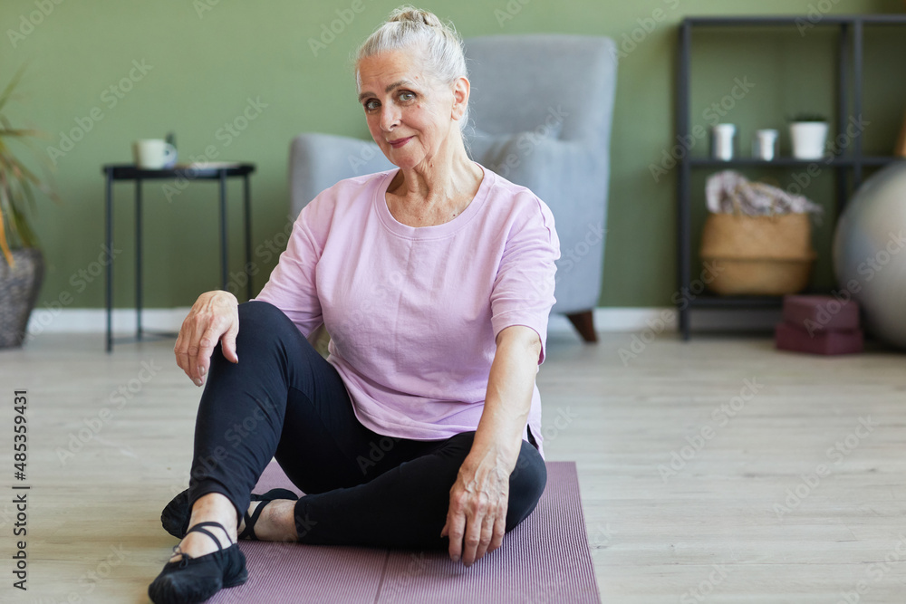 Happy active senior female in sports clothes looking at camera while sitting on yoga mat in living-room on background of furniture