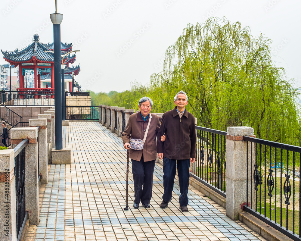 Senior Man and Woman Walking Outside. A senior couple, 80 years old, helping each other, arm in arm, is walking on a park, woman holding a walking stick.. Senior Man and Woman Walking Outside. A senior couple, 80 years old, helping each other, arm in arm, is walking on a park, woman holding a walking stick..