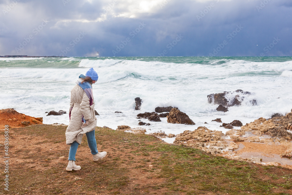 Stylish woman walks on the shore of the winter sea. She dressed in a beige coat and a hat, there was a storm on the sea
