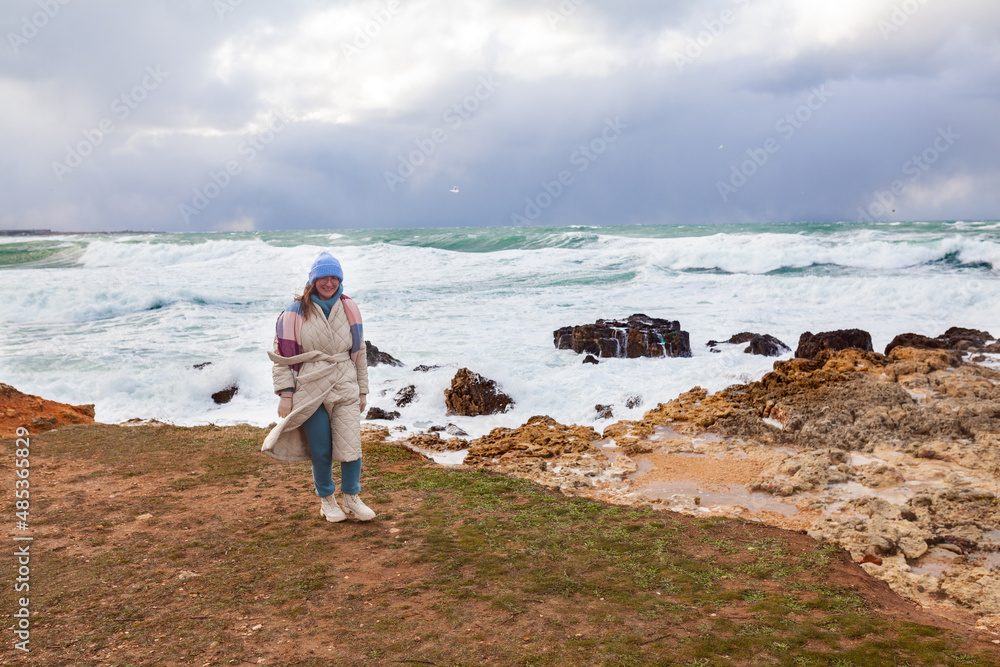 Stylish woman walks on the shore of the winter sea. She dressed in a beige coat and a hat, there was a storm on the sea