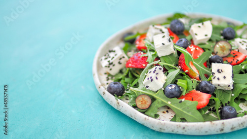 Salad with arugula, feta cheese and berries - strawberry, blueberry, in craft plate. Summer salad idea and recipe for healthy vegetarian lunch, dinner. Close up. Copy space. Colorful blue background.