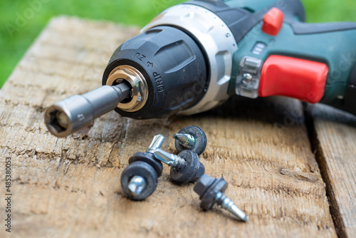 A close-up of an electric screwdriver with a nozzle on lies on a wooden background. Next to it, roofing screws on metal are laid out in a row. The concept of repair and construction