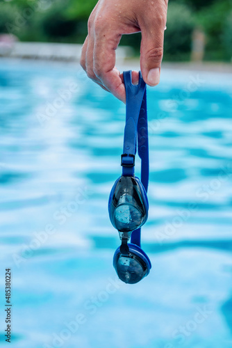 Man`s hand holding professional swimming goggles with pool blue water on background photo