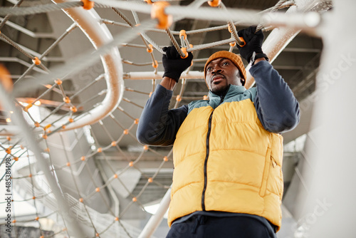 Young African American sportsman in warm sports clothes pulling up on rope net while exercising during outdoor workout photo