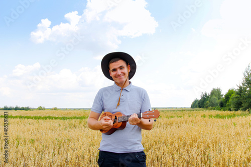 Singer. Young beautiful caucasian man playing ukulele outside. Handsome hipster guy plays ukulele guitar on nature background. Male in a cowboy hat smiles and plays on the guitar photo