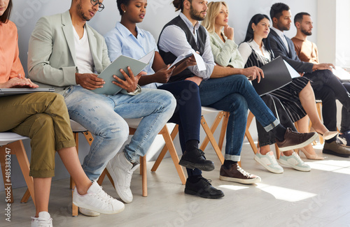 Crop image of diverse employees sit on chairs in row listen to seminar or training in office. Job candidates or applicants in line wait for interview for vacant position. Employment, hiring concept.