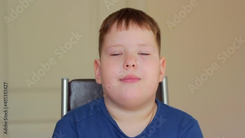 A fat boy sits on a chair and yawns. He's wearing a blue T-shirt.