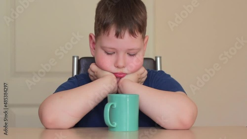 There is a green mug on the table. A fat child is sitting at the table, propping his chin on his hands, looking at a mug.