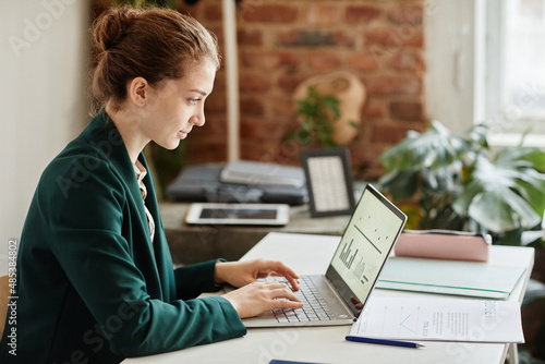 Young confident businesswoman in formal clothes typing on laptop keyboard while analyzing financial data on screen photo