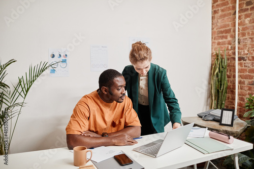 Young serious blackman looking at laptop screen while listening to his colleague explanations of financial data at meeting photo