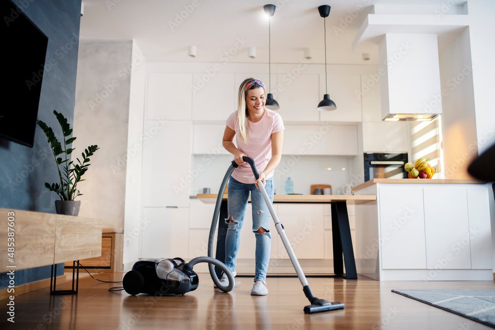 A smiling woman vacuuming living room floor at her home. - obrazy, fototapety, plakaty 