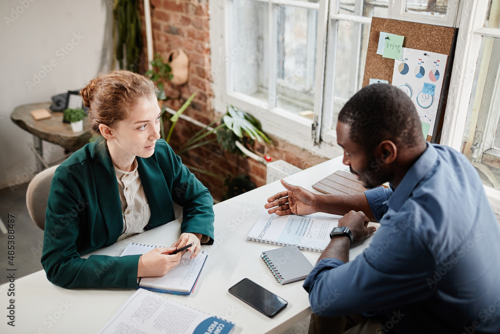 Two young confident intercultural colleagues with business documents discussing working plan or data at working meeting Two young confident intercultural colleagues with business documents discussing working plan or data at working meeting