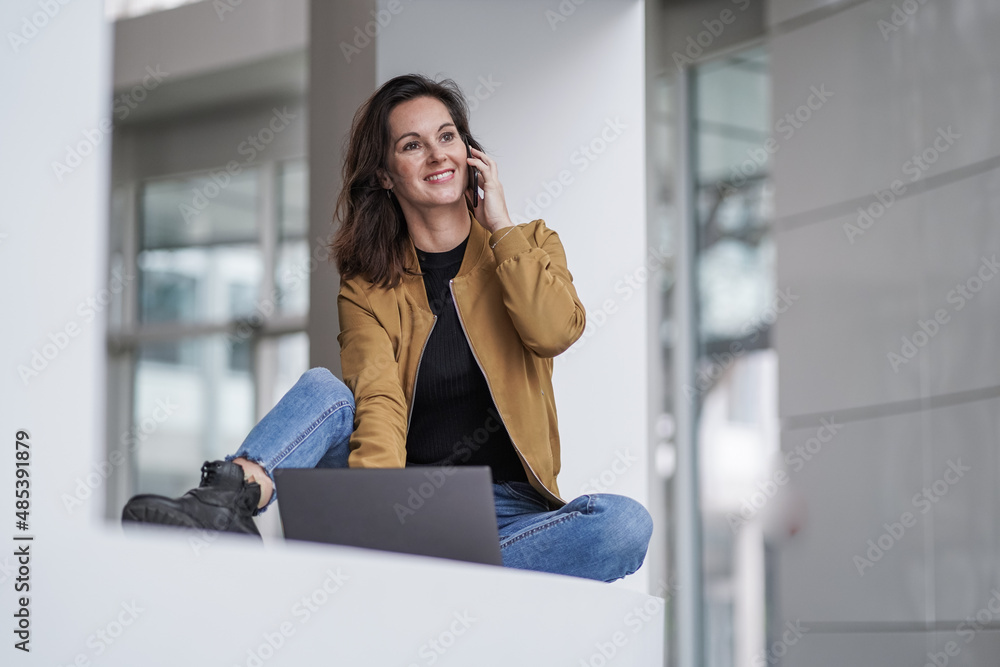 Fototapeta premium smiling easygoing remote working lady girl calling and chatting infront of a laptop or notebook in casual jeans outfit with her mobile phone in a modern hotel lobby on a white ground