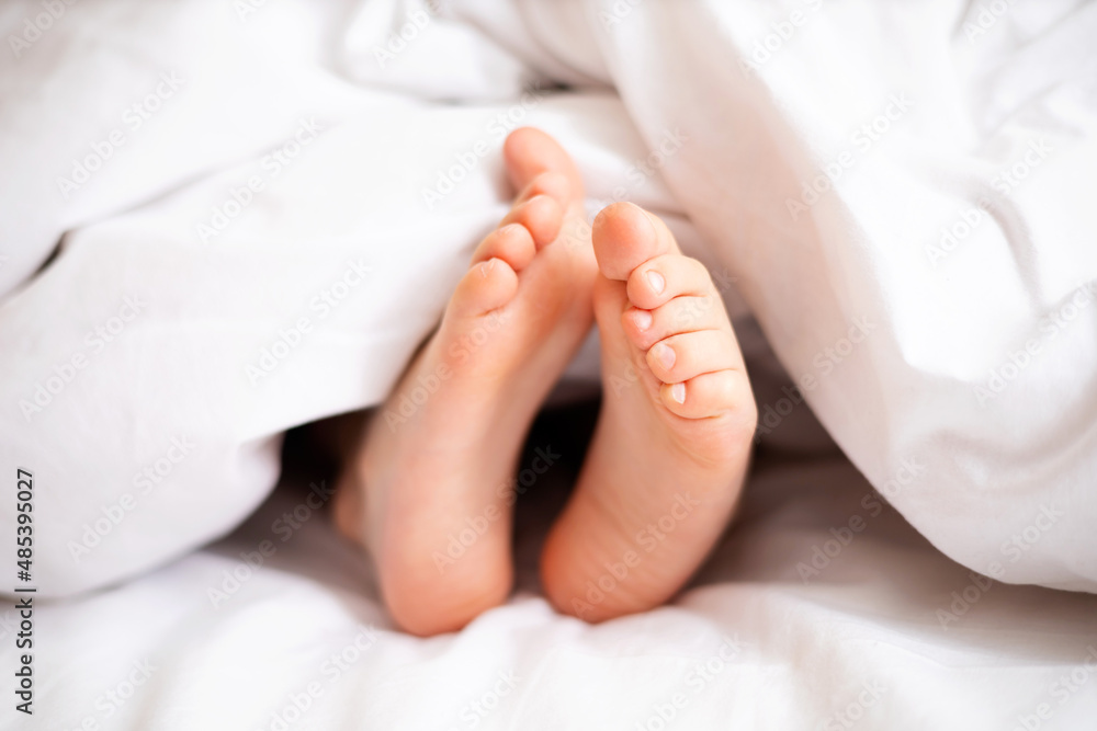 Children's legs stick out from under a white blanket in the bedroom. Dream. Morning. Lifestyle Children's legs stick out from under a white blanket in the bedroom. Dream. Morning. Lifestyle