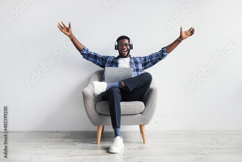 Excited African American guy in headphones greeting someone while chatting online on laptop, sitting in armchair