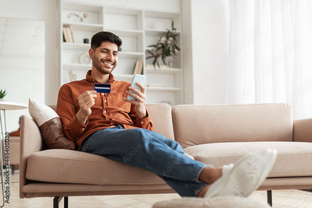 Obraz premium Smiling Arab guy using phone and credit card at home