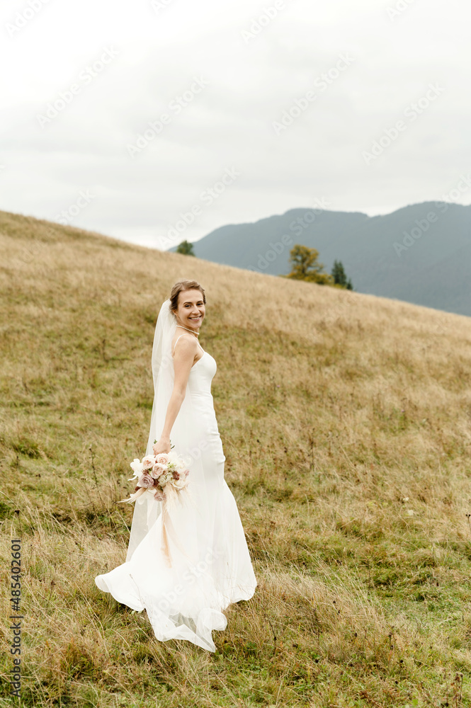 Bride portrait. A young girl in a white wedding dress and with a bouquet of flowers and greenery in her hands against the backdrop of mountains and forests at sunset